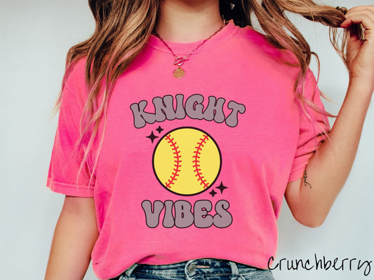 A woman wearing a cute crunchberry colored shirt with gray text saying Knight Vibes. There is a yellow baseball with red stitching with black twinkling stars around it between the text.