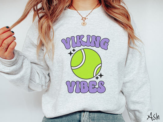 A woman wearing a cute ash colored sweatshirt with purple text saying Viking Vibes. There is a neon tennis ball with white stripes and black twinkling stars around it between the text.