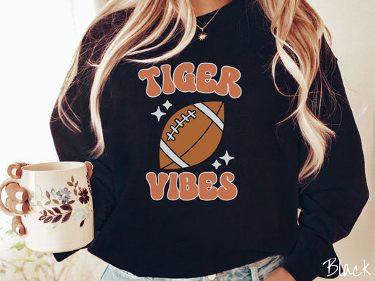 A woman wearing a cute black colored sweatshirt with orange text saying Tiger Vibes. There is a brown football with white twinkling stars around it between the text.