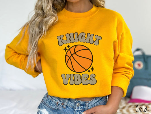 A woman wearing a cute gold colored sweatshirt with gray text saying Knight Vibes. There is an orange basketball with black twinkling stars around it between the text.