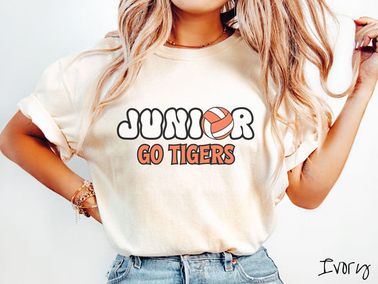 A woman wearing a cute ivory colored t-shirt with the word Junior in black and white font, the O in Junior is an orange and white volleyball. Below Junior are the words in orange and black font Go Tigers.