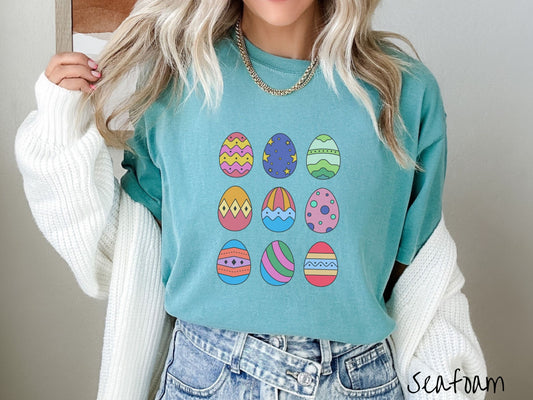 A woman wearing a cute, vintage seafoam colored shirt with a three by three grid of colorful Easter eggs. One is pink, red, and yellow, another blue with stars, and another is different shades of green next to five more egg designs.