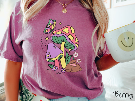 A woman wearing a cute, vintage berry colored shirt with a purple mushroom and a pink and yellow mushroom together with a brown bunny rabbit laying on the ground by purple and white flowers, and a colorful butterfly is fluttering above it all.