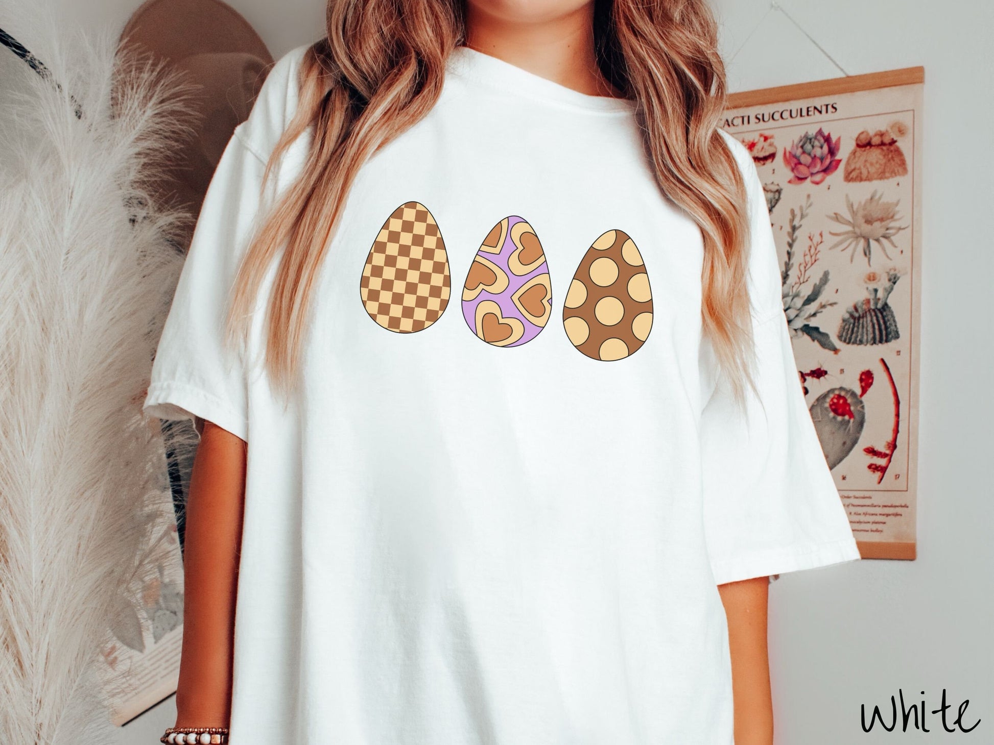 A woman wearing a cute, vintage white colored shirt with three Easter eggs. The left egg has a light yellow and brown checkerboard pattern, the middle is purple with light yellow brown hearts, and the right is brown with light yellow circles.