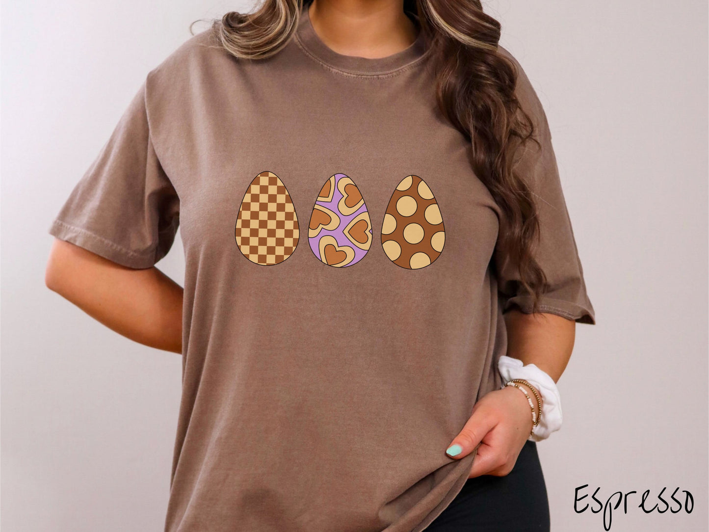 A woman wearing a cute, vintage espresso colored shirt with three Easter eggs. The left egg has a light yellow and brown checkerboard pattern, the middle is purple with light yellow brown hearts, and the right is brown with light yellow circles.