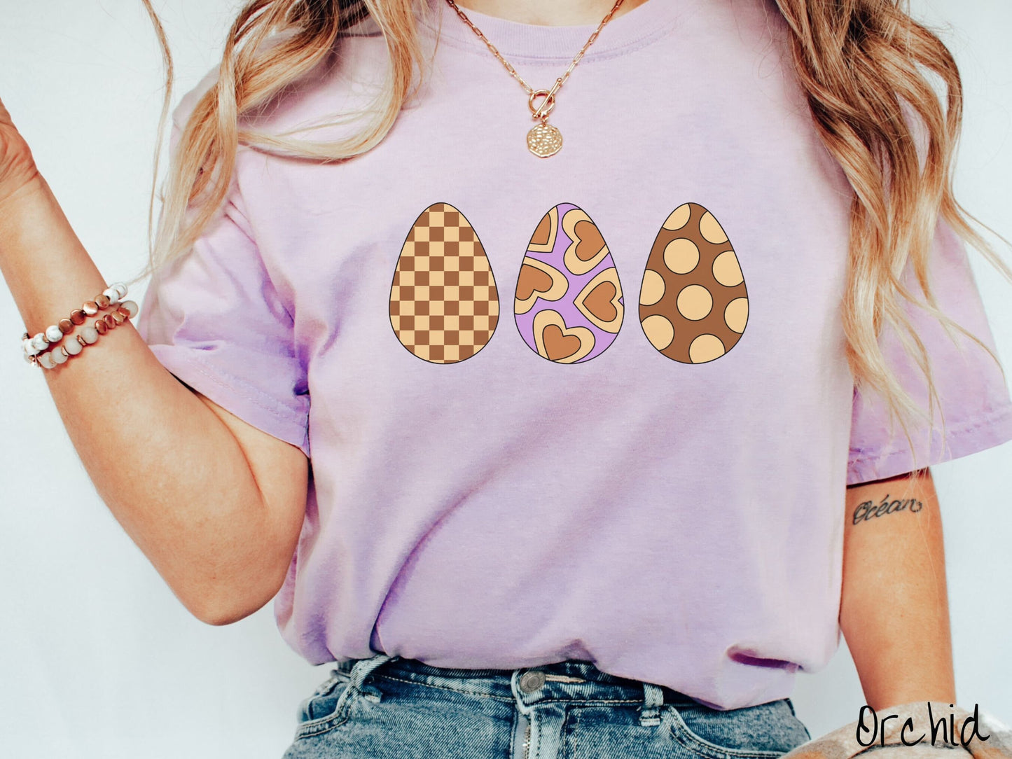 A woman wearing a cute, vintage orchid colored shirt with three Easter eggs. The left egg has a light yellow and brown checkerboard pattern, the middle is purple with light yellow brown hearts, and the right is brown with light yellow circles.