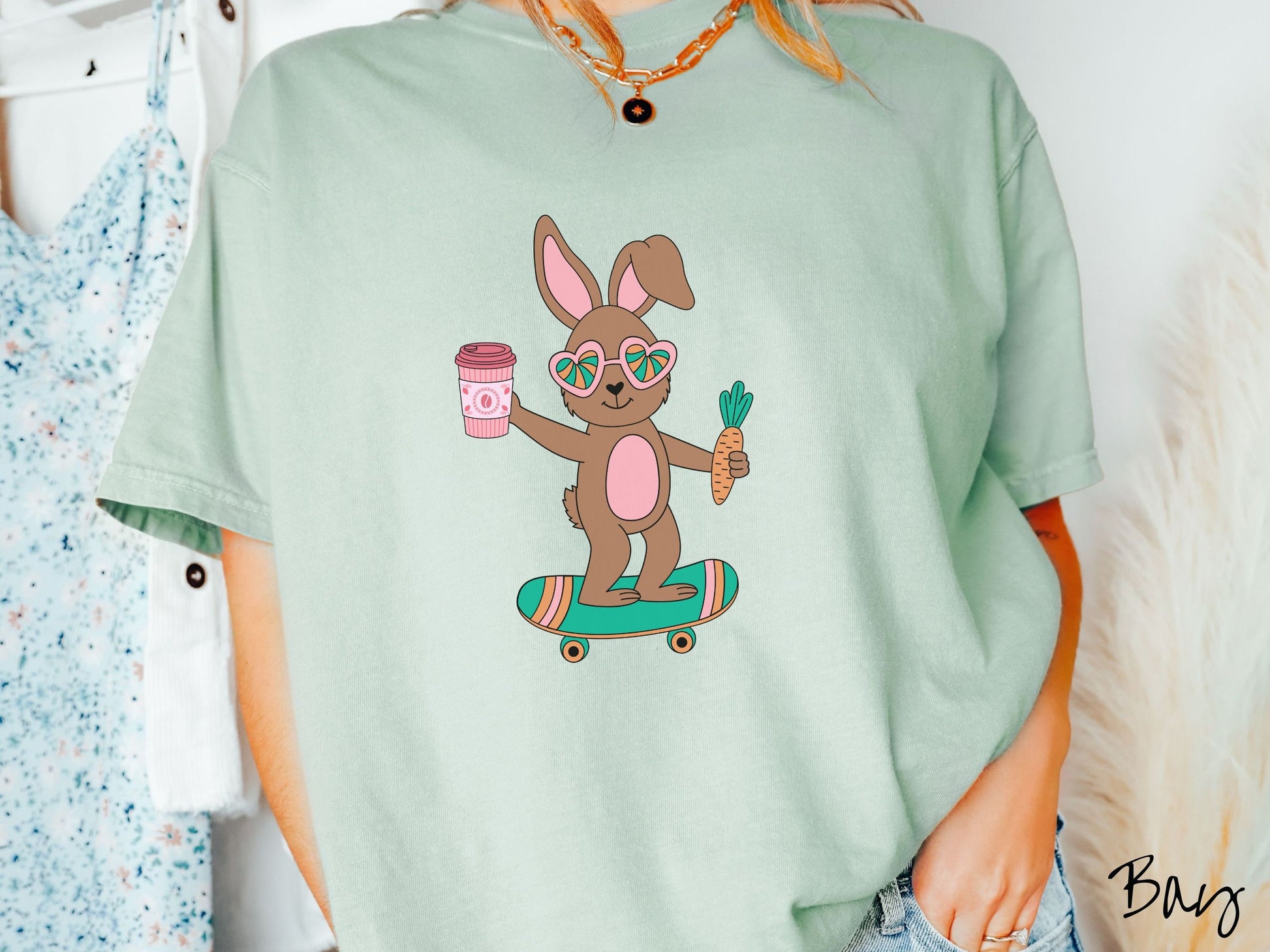 A woman wearing a cute, vintage bay colored shirt with a brown bunny rabbit with heart-shaped glasses, a carrot in one hand, and a pink to-go coffee in the other, riding a green, pink, and orange skateboard.