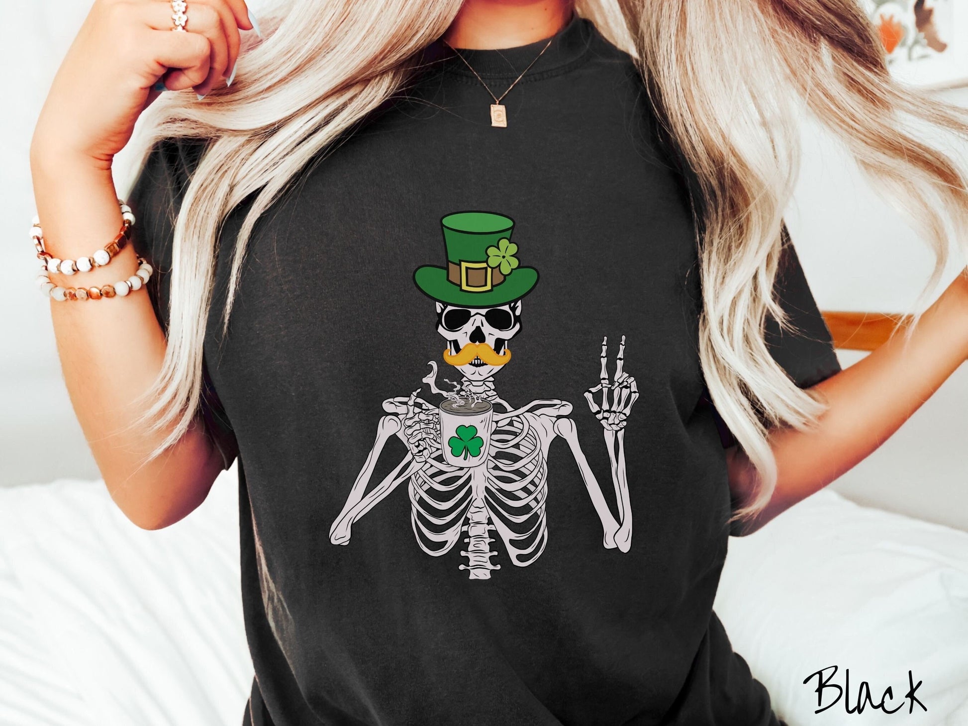 A woman wearing a vintage, black colored shirt with a skeleton wearing a green top hat with a green clover and an orange mustache showing the peace sign with its left hand and drinking out of a steaming hot coffee cup with a three leaf clover on it.