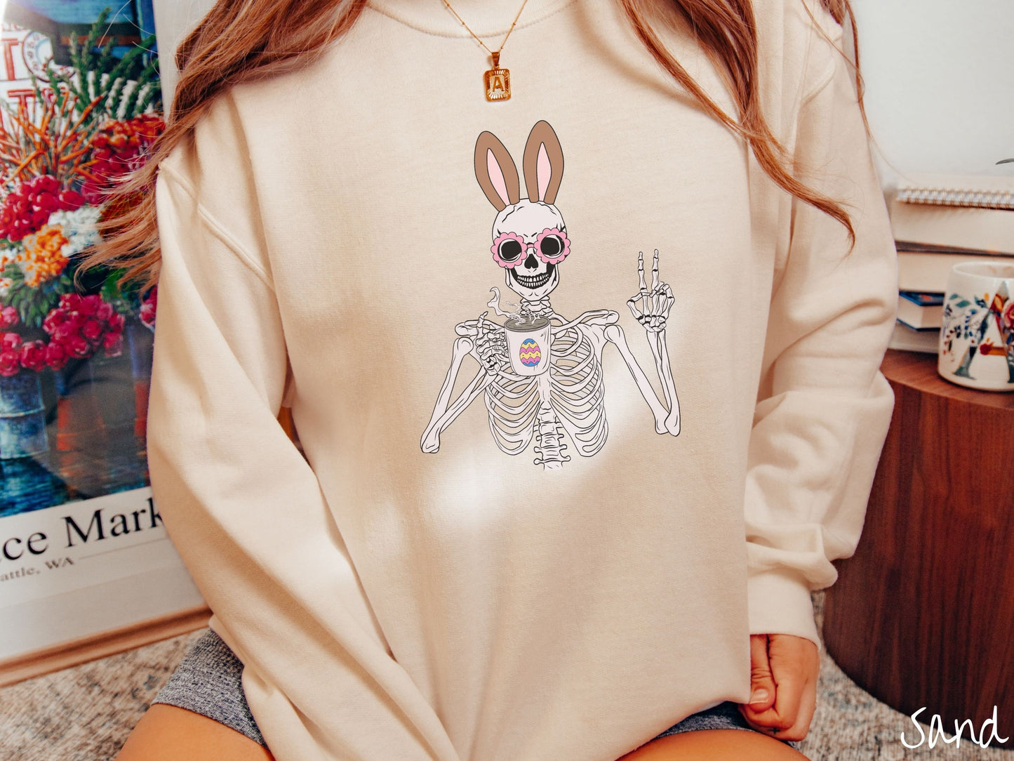 A woman wearing a cute, vintage sand colored sweatshirt with a pink-toned skeleton wearing pink glasses and brown bunny ears, holding a cup of coffee and holding up a peace sign with the other hand. The white coffee cup has a colorful Easter egg.