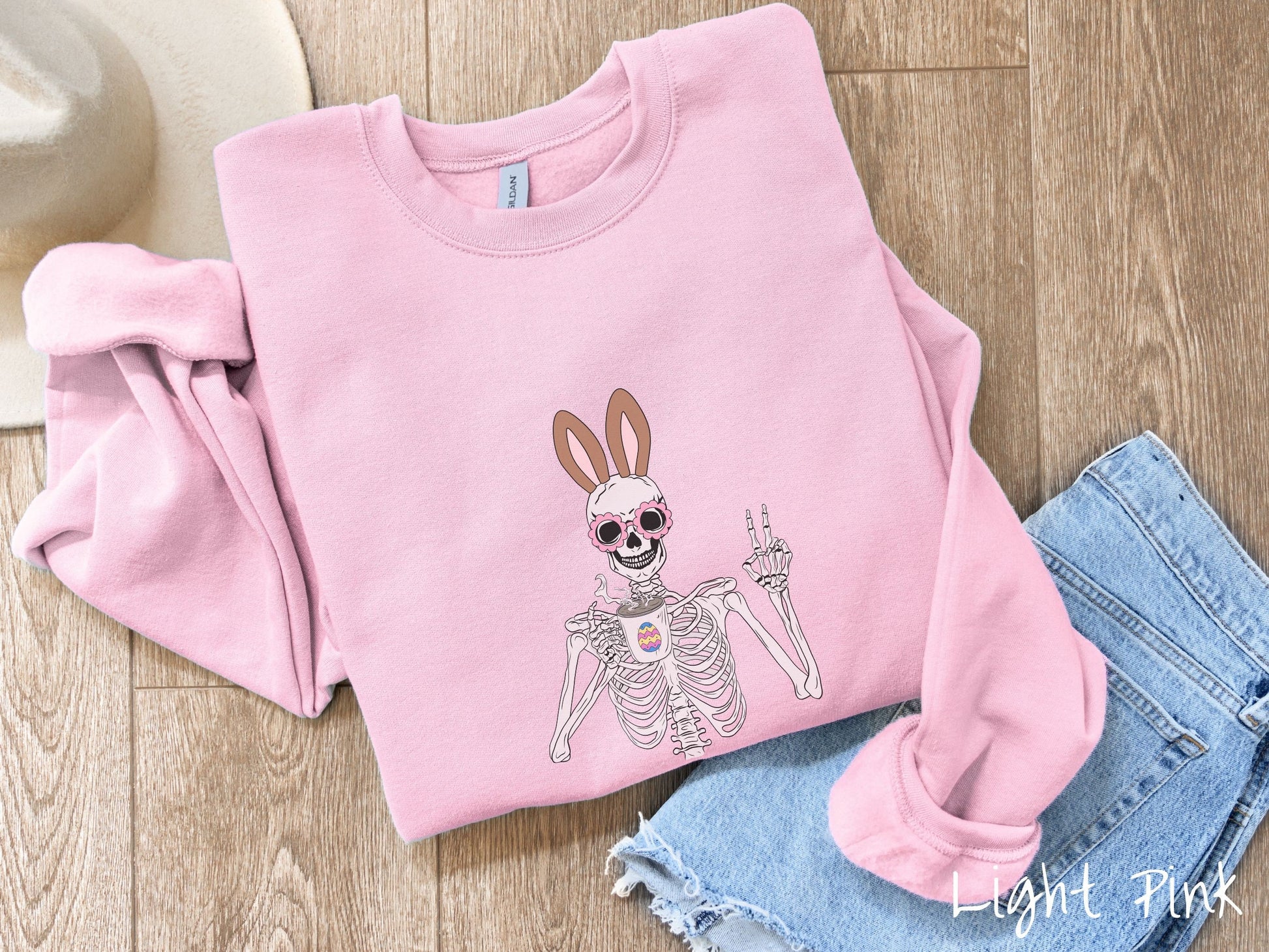 A cute, vintage light pink colored sweatshirt with a pink-toned skeleton wearing pink glasses and brown bunny ears, holding a cup of coffee and holding up a peace sign with the other hand. The white coffee cup has a colorful Easter egg.