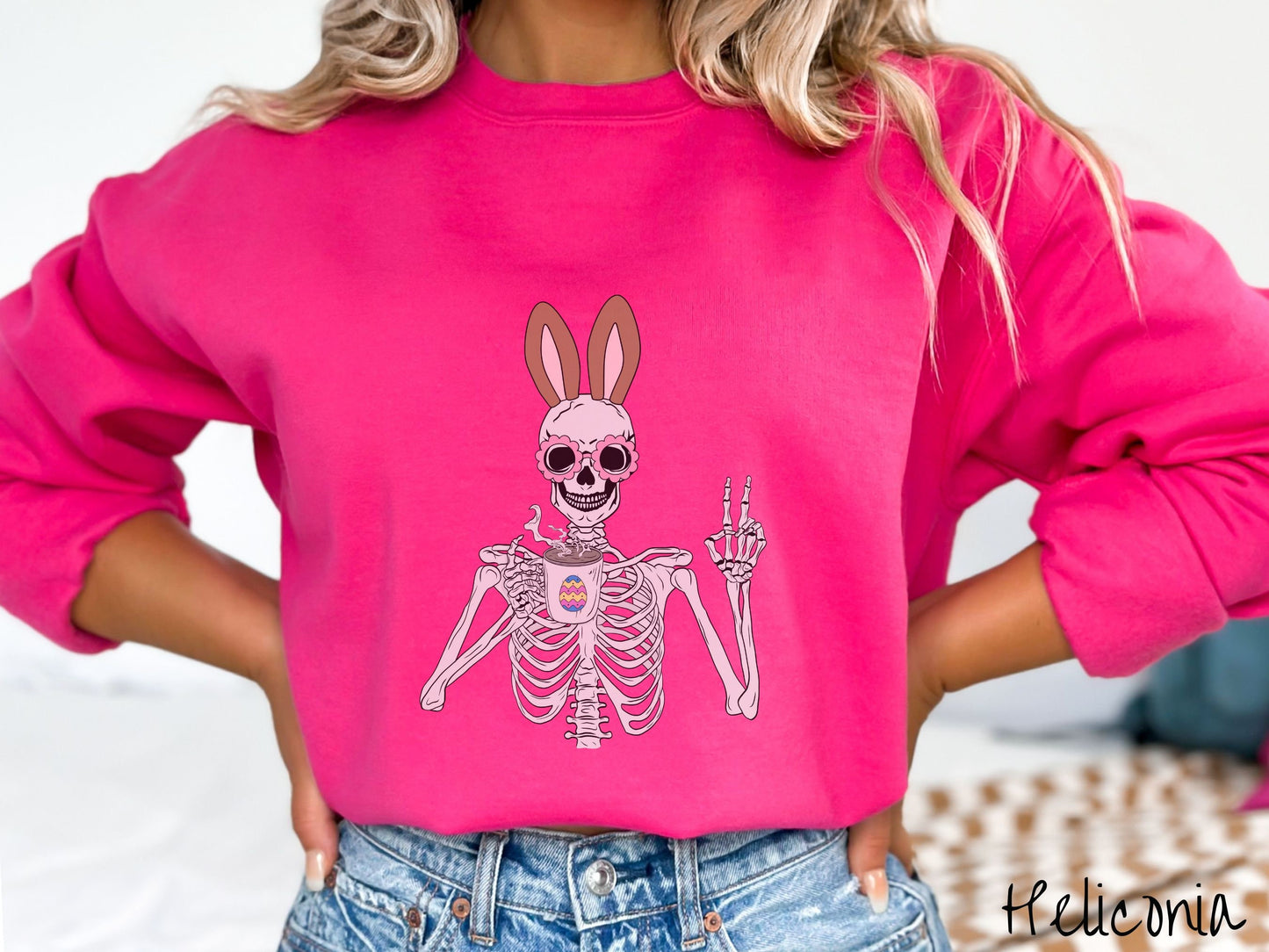 A woman wearing a cute, vintage heliconia colored sweatshirt with a pink-toned skeleton wearing pink glasses and brown bunny ears, holding a cup of coffee and holding up a peace sign with the other hand. The coffee cup has a colorful Easter egg.