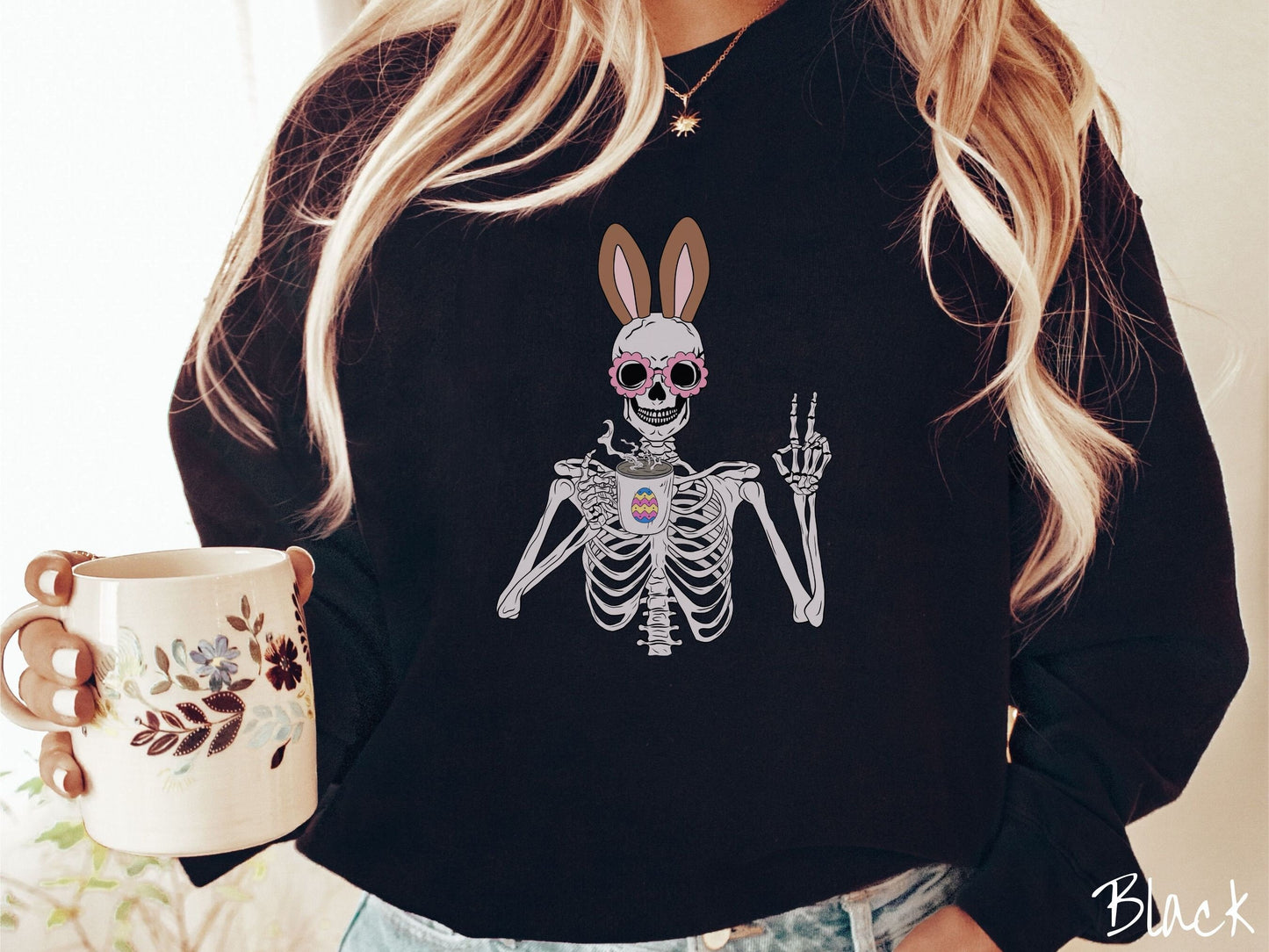 A woman wearing a cute, vintage black colored sweatshirt with a pink-toned skeleton wearing pink glasses and brown bunny ears, holding a cup of coffee and holding up a peace sign with the other hand. The white coffee cup has a colorful Easter egg.