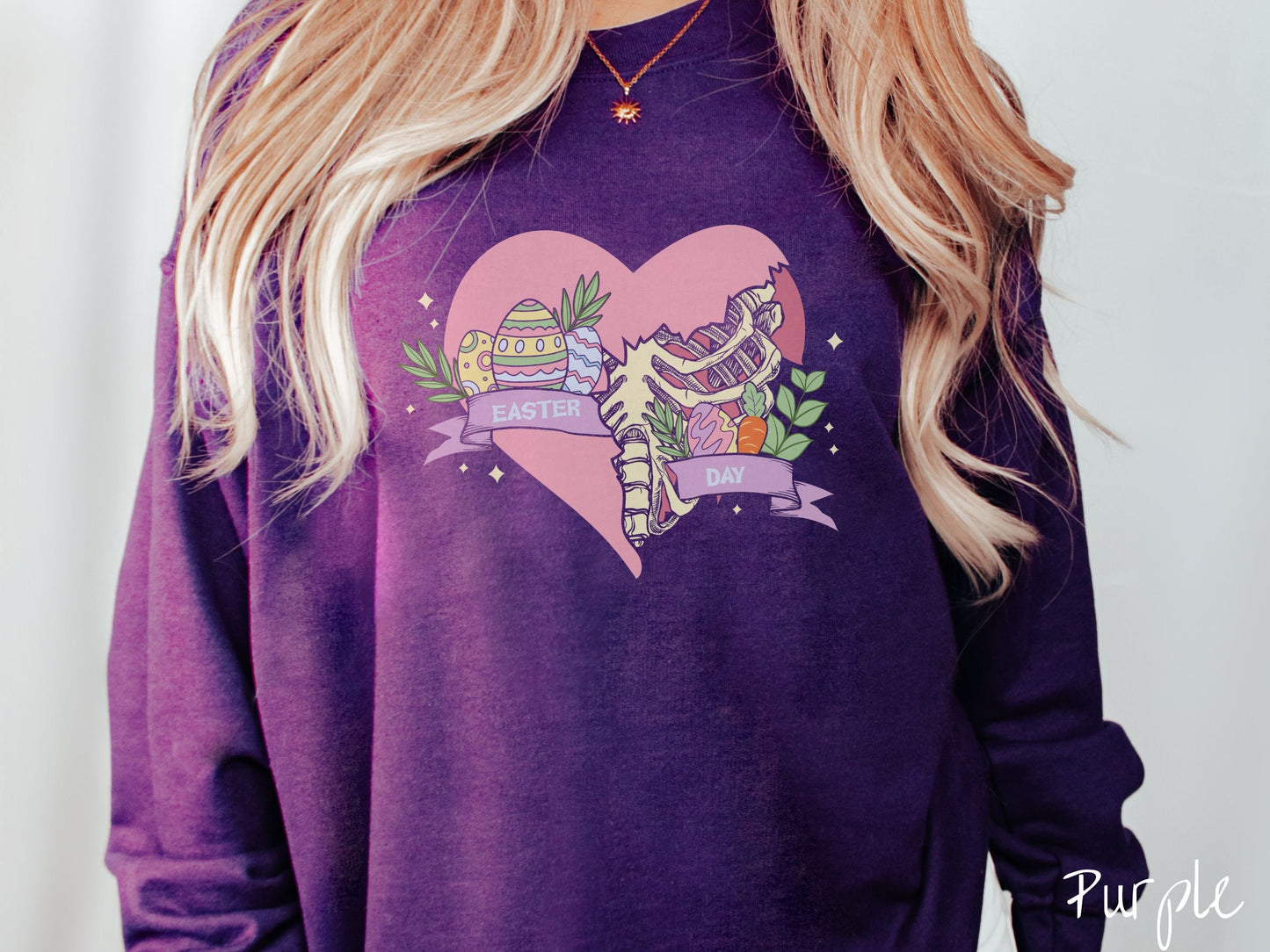 A woman wearing a cute, vintage purple colored sweatshirt with a pink heart that is cracking and underneath is a skeleton rib cage. There is a pink banner across the heart that says Easter Day, and above that are colorful Easter eggs and leaves.