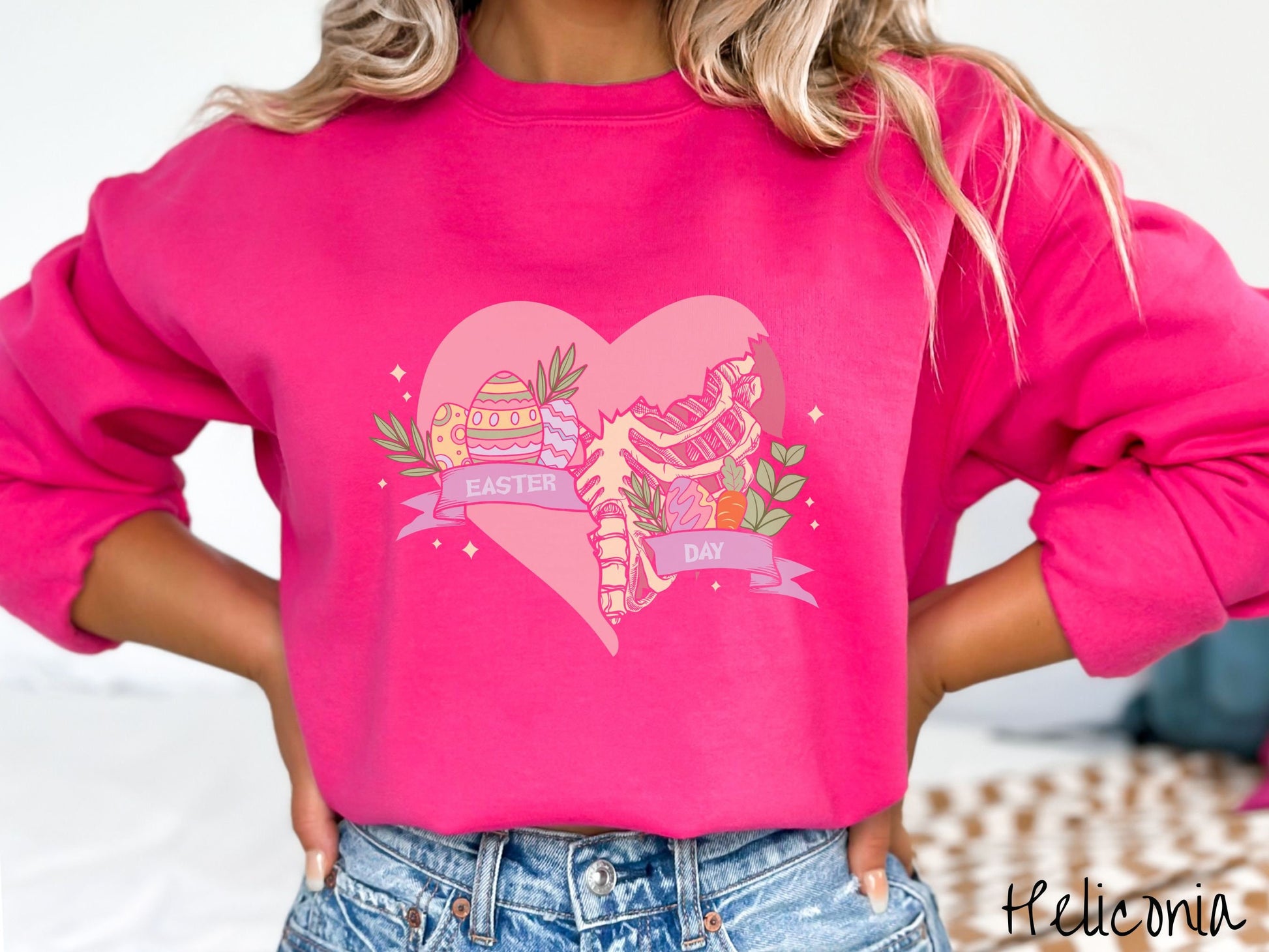 A woman wearing a cute, vintage heliconia colored sweatshirt with a pink heart that is cracking and underneath is a skeleton rib cage. There is a pink banner across the heart that says Easter Day, and above that are colorful Easter eggs and leaves.
