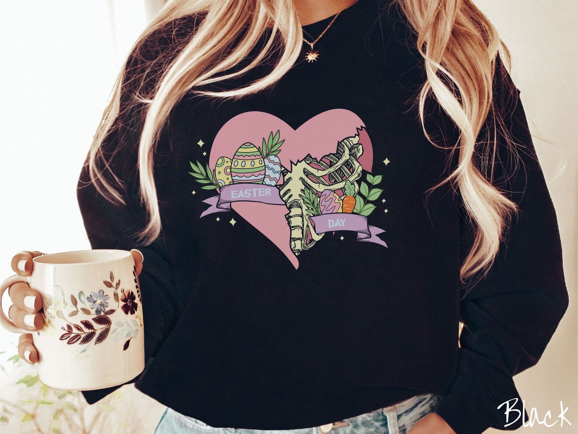 A woman wearing a cute, vintage black colored sweatshirt with a pink heart that is cracking and underneath is a skeleton rib cage. There is a pink banner across the heart that says Easter Day, and above that are colorful Easter eggs and leaves.