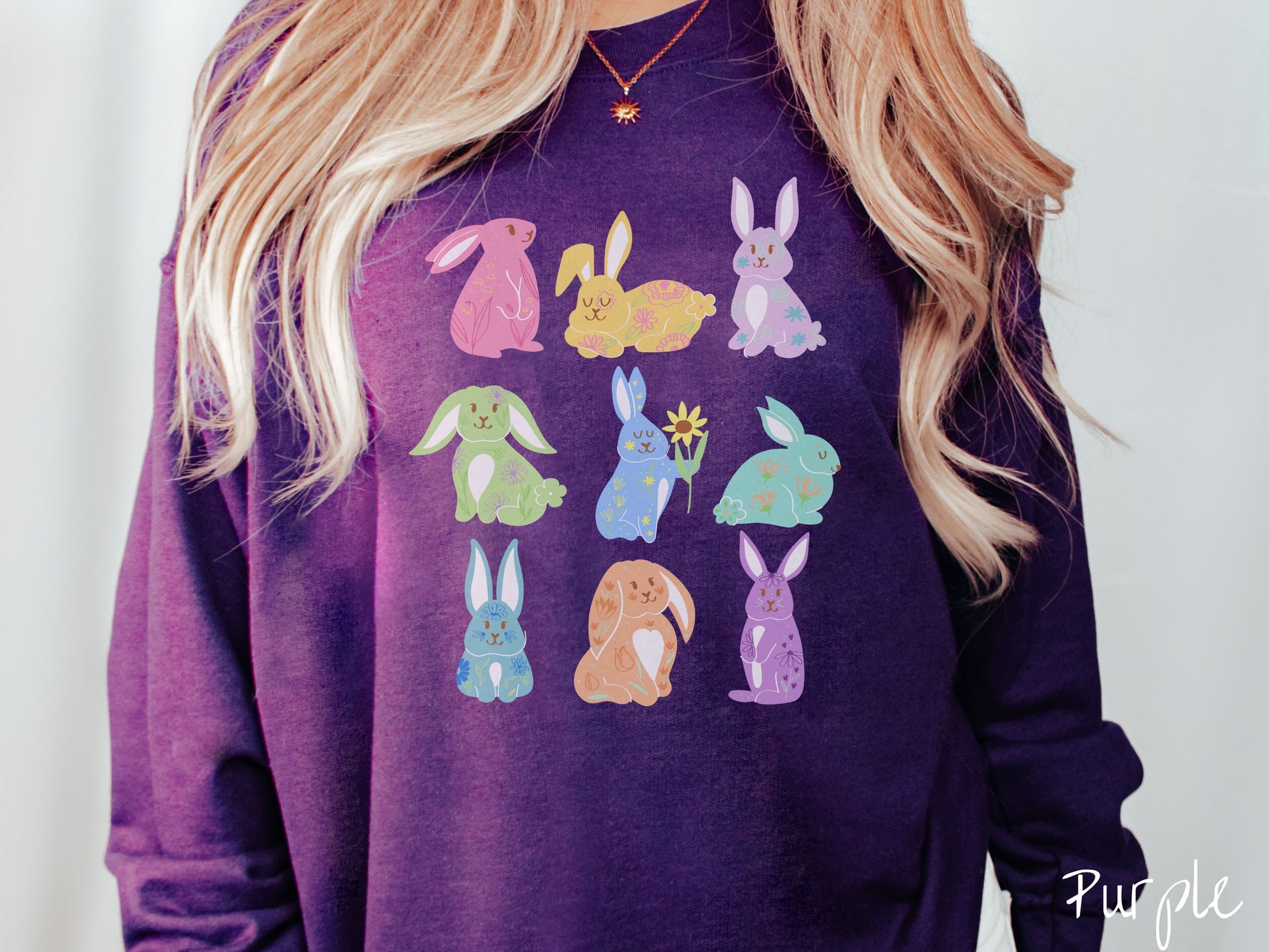 A woman wearing a cute, vintage purple colored sweatshirt with a three by three grid of colorful bunny rabbits. They are from left to right pink, yellow, purple, green, blue, seafoam, light blue, orange, and purple with flowers near them.