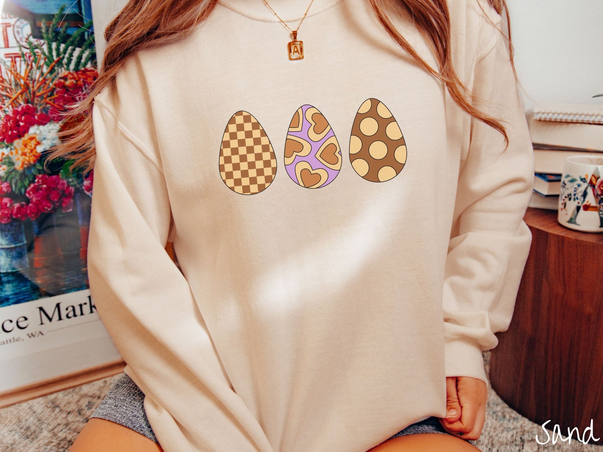 A woman wearing a cute, vintage sand colored sweatshirt with three Easter eggs. The left has a light yellow and brown checkerboard pattern, the middle is purple with light yellow brown hearts, and the right is brown with light yellow circles.