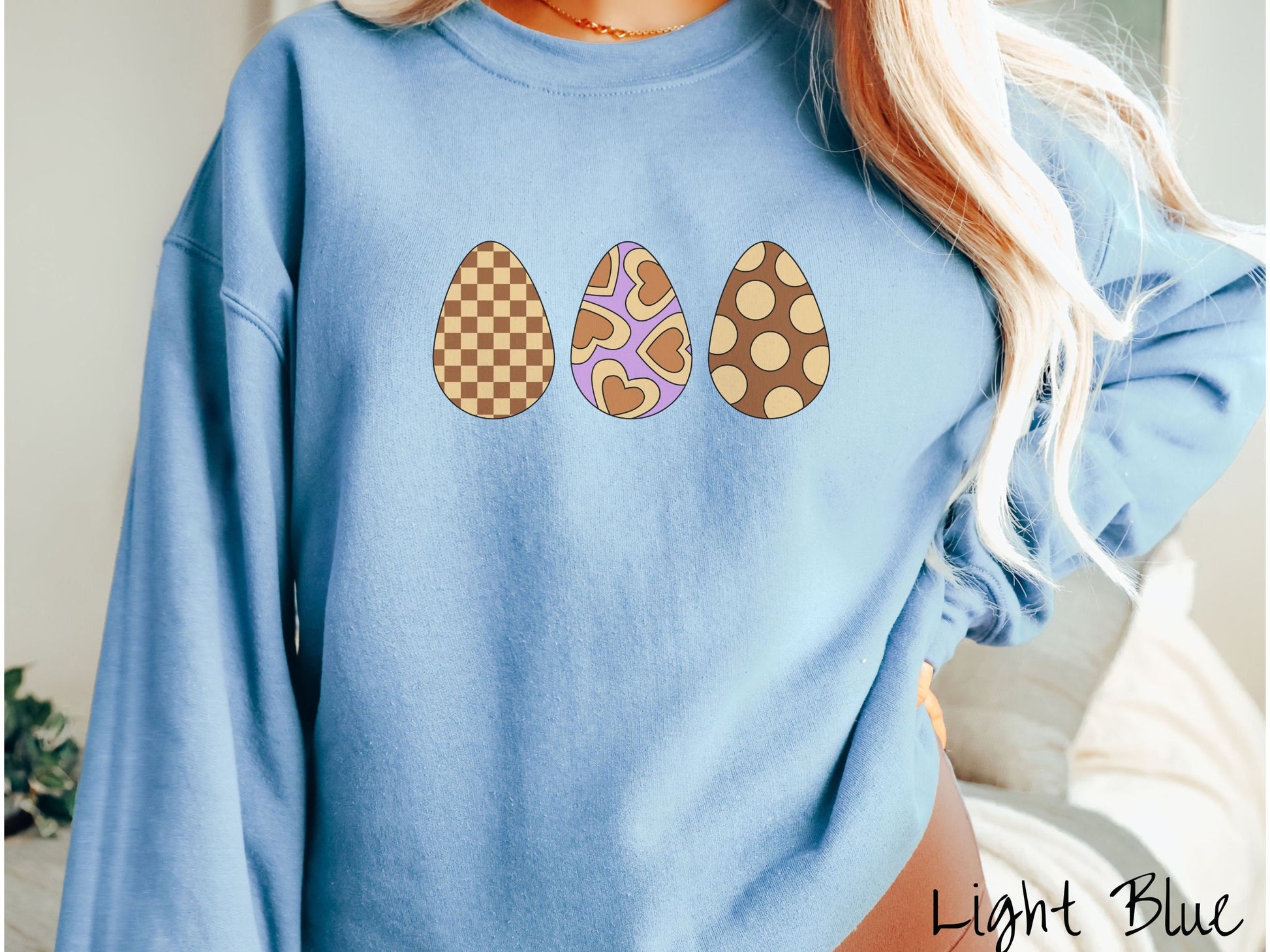 A woman wearing a cute, vintage light blue colored sweatshirt with three Easter eggs. The left has a light yellow and brown checkerboard pattern, the middle is purple with light yellow brown hearts, and the right is brown with light yellow circles.