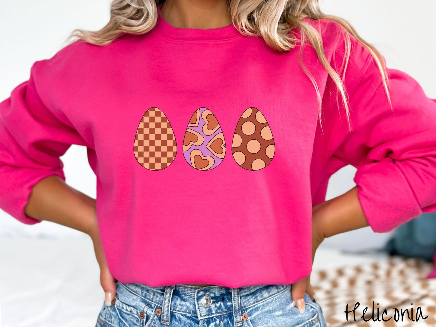 A woman wearing a cute, vintage heliconia colored sweatshirt with three Easter eggs. The left has a light yellow and brown checkerboard pattern, the middle is purple with light yellow brown hearts, and the right is brown with light yellow circles.