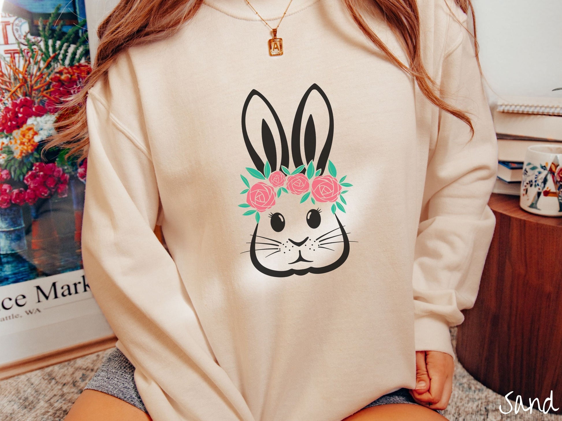 A woman wearing a cute, vintage sand colored sweatshirt with a black outline of a bunny rabbit with eyelashes, whiskers, and floppy ears that has a pink flowered headband resting below its large ears.