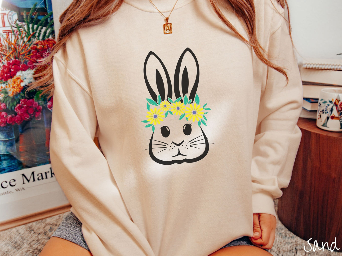A woman wearing a cute, vintage sand colored sweatshirt with a black outline of a bunny rabbit with eyelashes and whiskers that has a yellow and purple flower headband resting below its large ears.