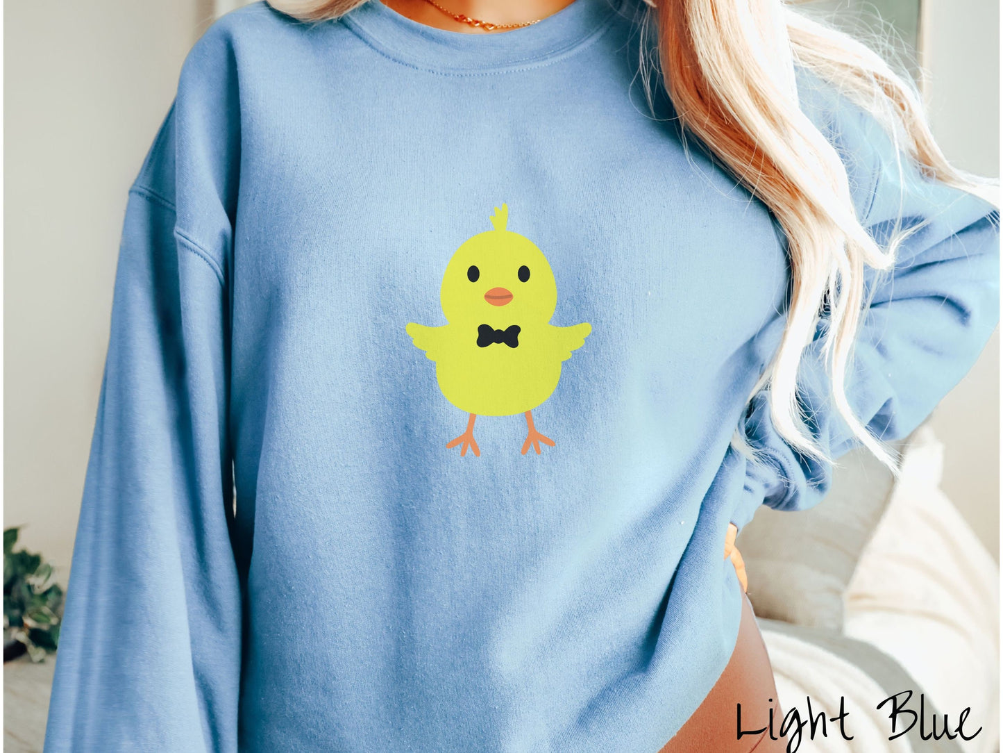 A woman wearing a cute, vintage light blue colored sweatshirt with a yellow baby chick with a black bowtie and its wings spread is standing and smiling. It has an orange mouth and feet and yellow feathers sticking out the top of its head.