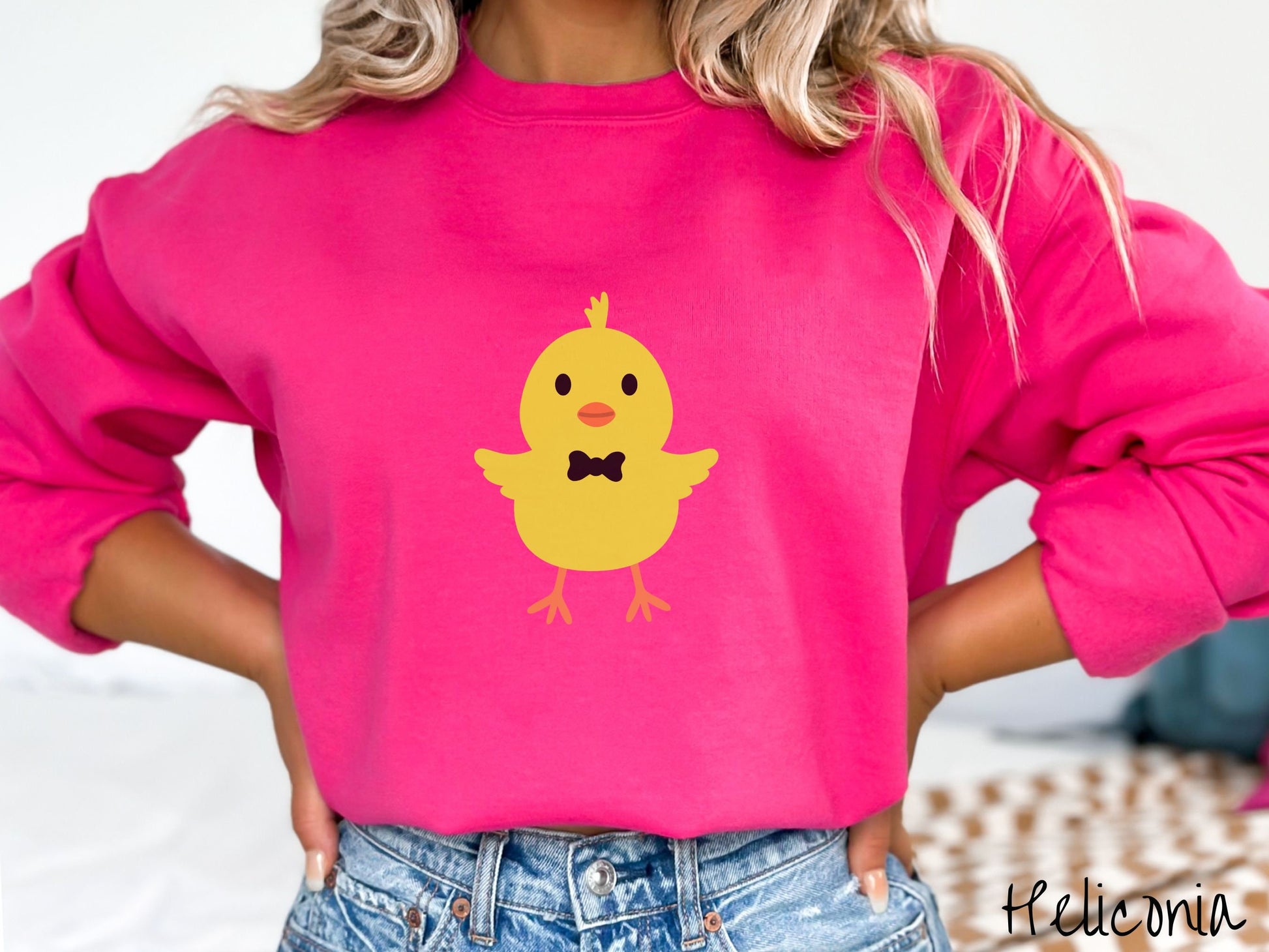 A woman wearing a cute, vintage heliconia colored sweatshirt with a yellow baby chick with a black bowtie and its wings spread is standing and smiling. It has an orange mouth and feet and yellow feathers sticking out the top of its head.