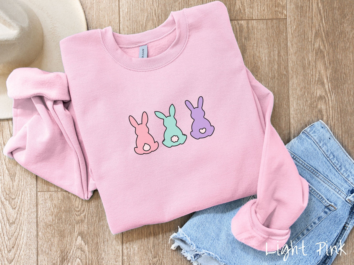 A cute, vintage light pink colored shirt with three colorful bunny rabbits sitting facing away. One is pink with a white tail, another bay with white tail, and the third is purple with a white, heart-shaped tail.