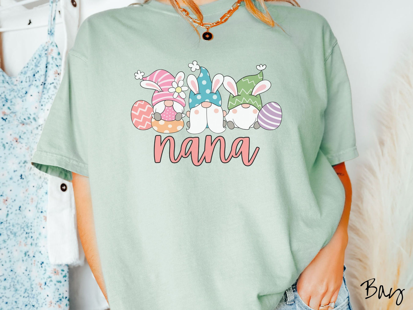 A woman wearing a cute, vintage bay colored shirt with text Nana below three gnomes with rabbit ears sitting next to each other. The left one is wearing pink, the middle blue, and right in green, with colorful easter eggs scattered about.