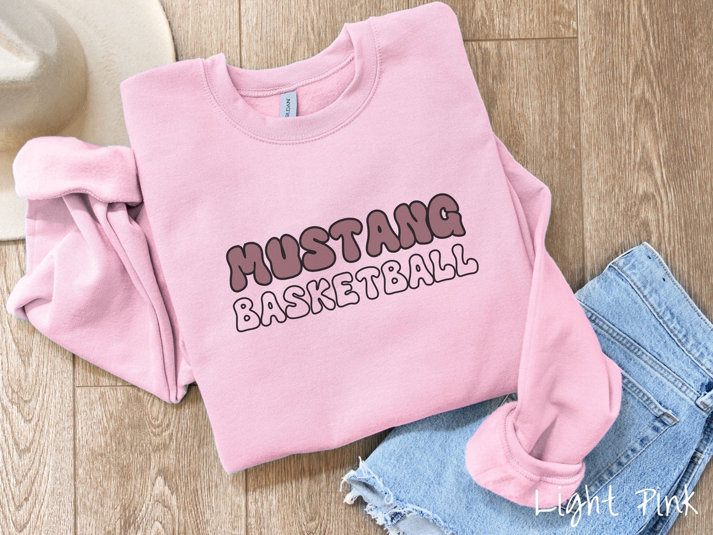 A cute, vintage light pink colored sweatshirt with the word Mustang across the front in purple, uppercase font. Below that is the word basketball in pink, uppercase font.