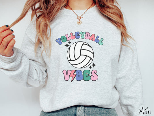 A woman wearing a cute, vintage ash colored sweatshirt with the word Volleyball across the front in colorful, uppercase fonts. Below that is a white volleyball and below that is the word vibes in colorful, uppercase fonts.