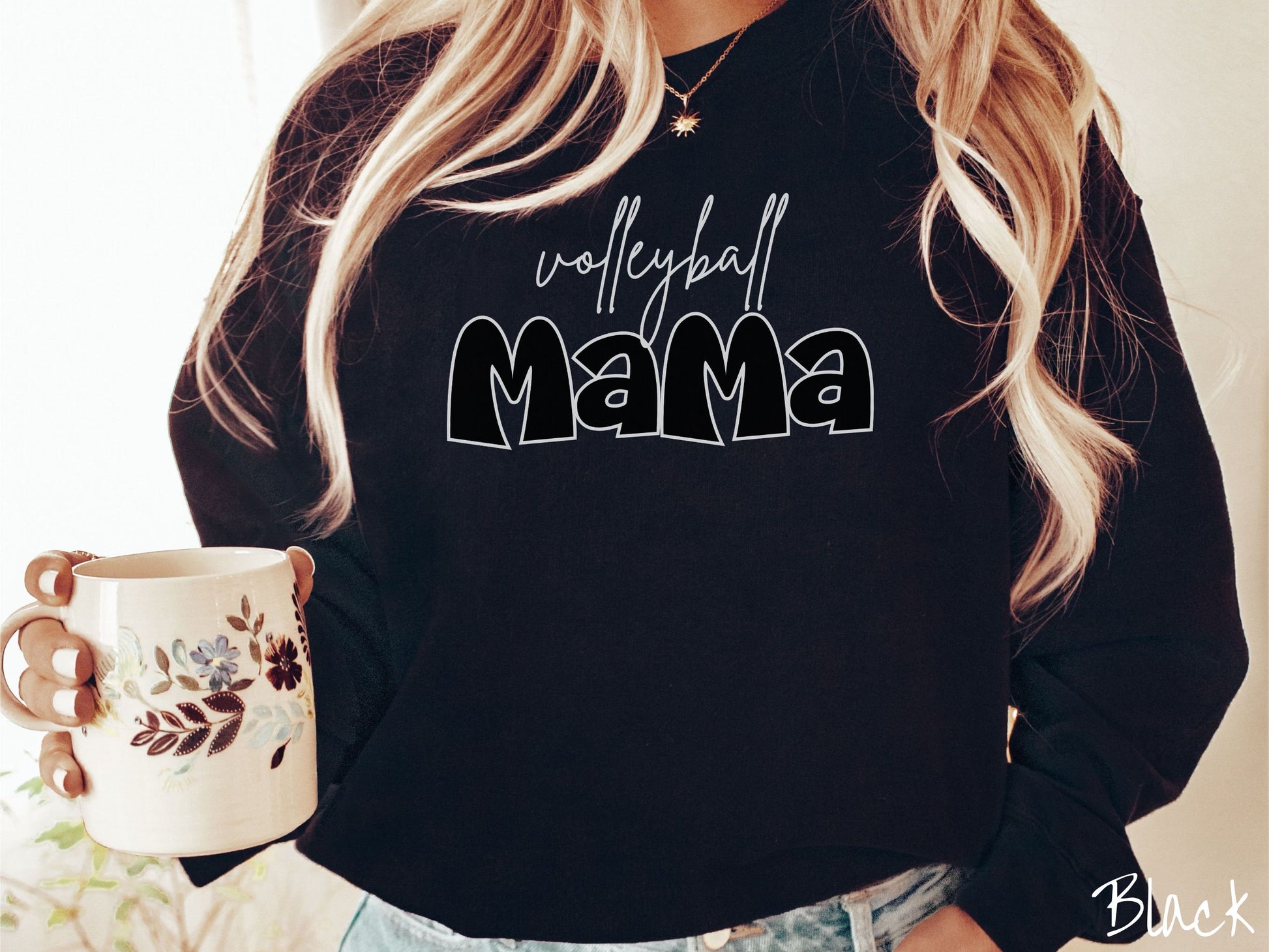 A woman wearing a cute, vintage black colored sweatshirt with the word volleyball across the front in white, undercase cursive writing. Below that is the word Mama in large, black font.