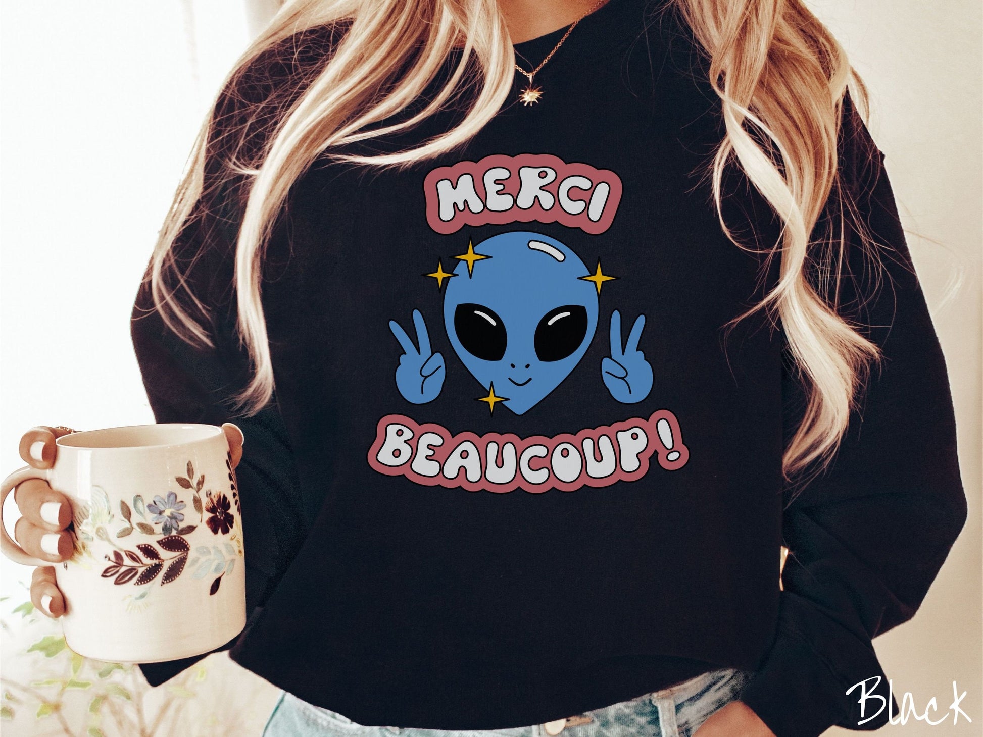 A woman wearing a cute, vintage black colored comfy sweatshirt with the text Merci Beaucoup! in white and maroon colored font. In between that is a blue alien with large black eyes smiling and holding up two peace signs.