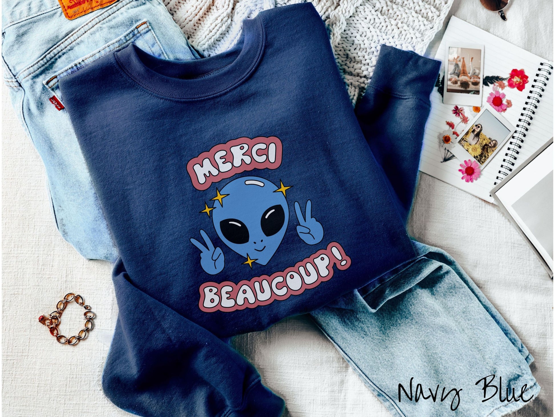 A cute, vintage navy blue colored comfy sweatshirt with the text Merci Beaucoup! in white and maroon colored font. In between that is a blue alien with large black eyes smiling and holding up two peace signs.