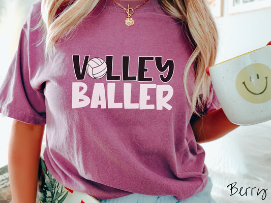 A woman wearing a cute, vintage berry colored Comfort Colors shirt with the word Volley across the front in black, uppercase font. The O in Volley is a white volleyball. Below that is the word Baller in white, uppercase font.