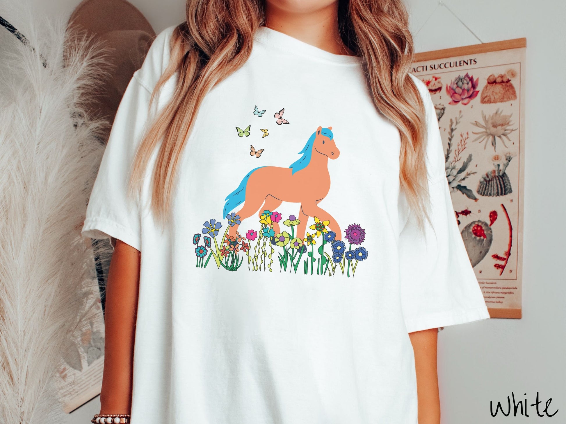 A woman wearing a cute, vintage white colored Comfort Colors shirt with a brown horse with blue hair walking through a field of colorful flowers and grass. There are colorful butterflies flying above the horse.