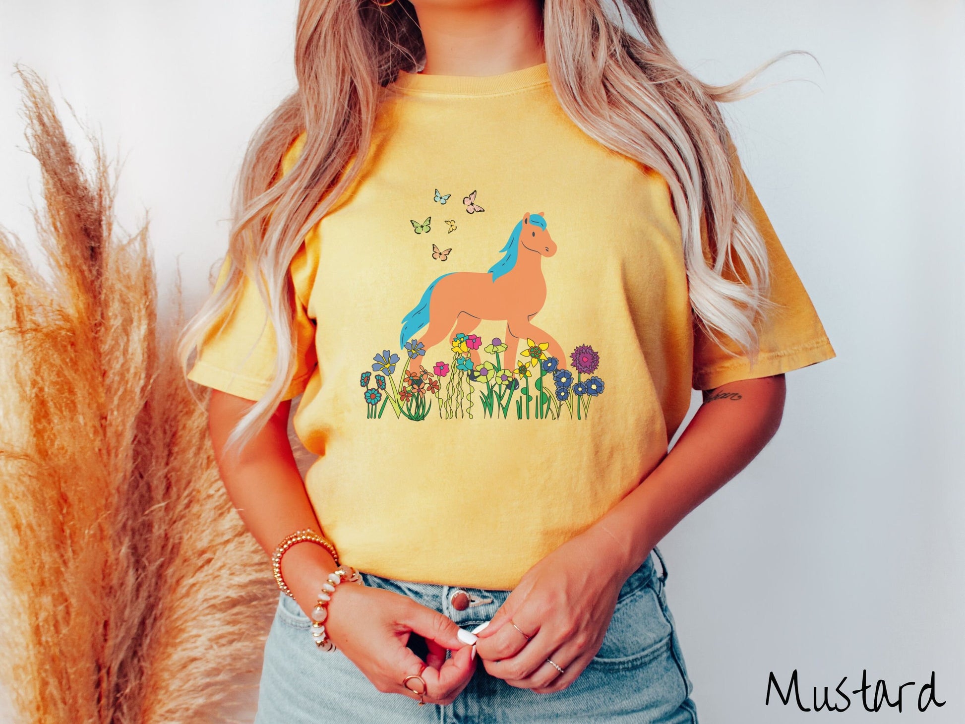 A woman wearing a cute, vintage gold colored Comfort Colors shirt with a brown horse with blue hair walking through a field of colorful flowers and grass. There are colorful butterflies flying above the horse.