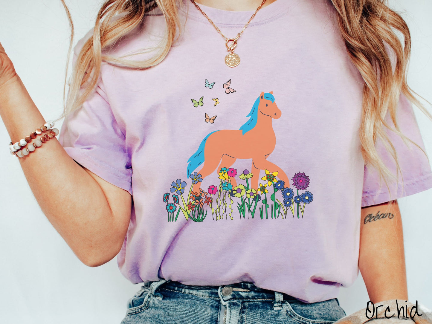 A woman wearing a cute, vintage orchid colored Comfort Colors shirt with a brown horse with blue hair walking through a field of colorful flowers and grass. There are colorful butterflies flying above the horse.