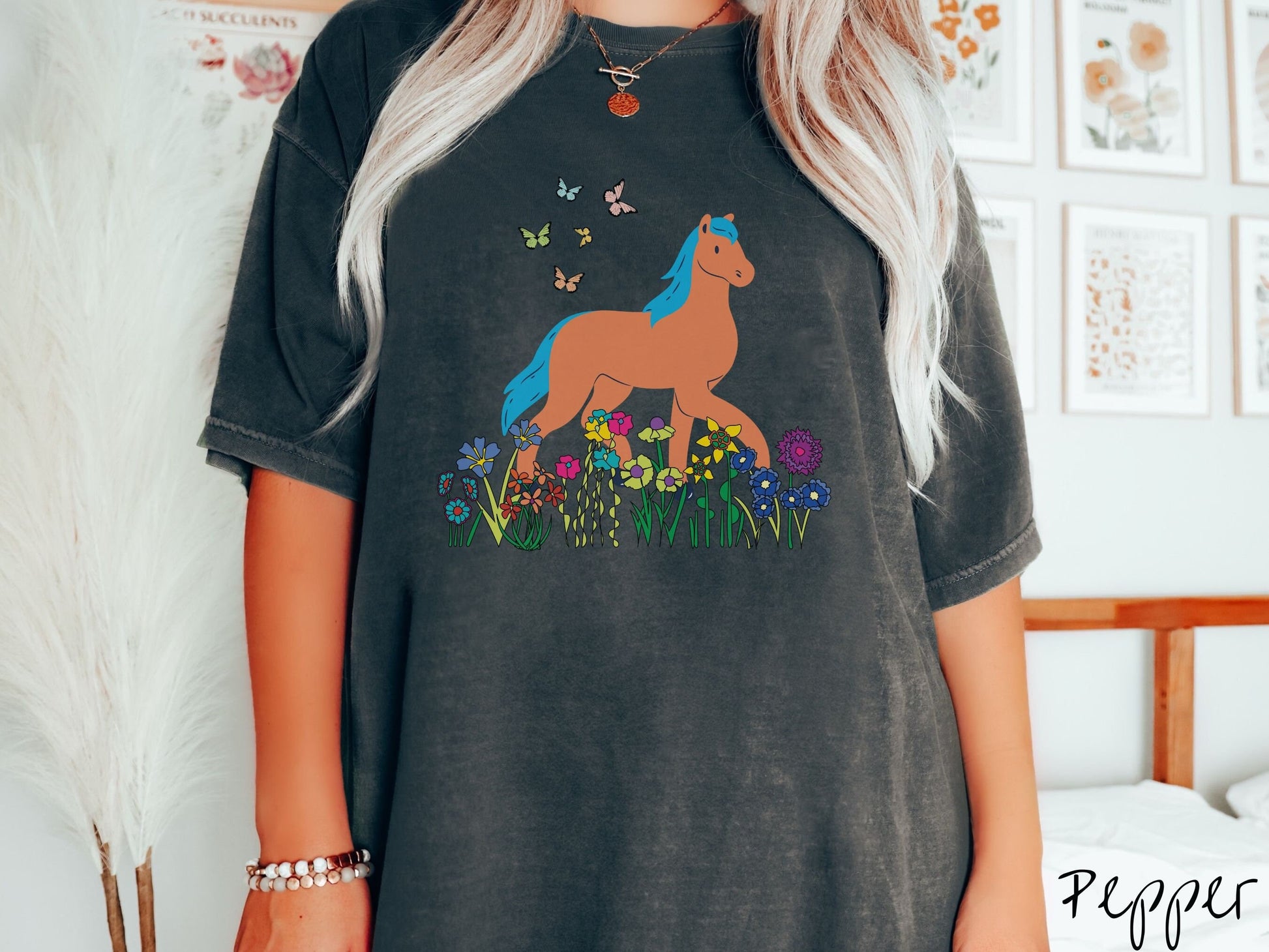 A woman wearing a cute, vintage pepper colored Comfort Colors shirt with a brown horse with blue hair walking through a field of colorful flowers and grass. There are colorful butterflies flying above the horse.