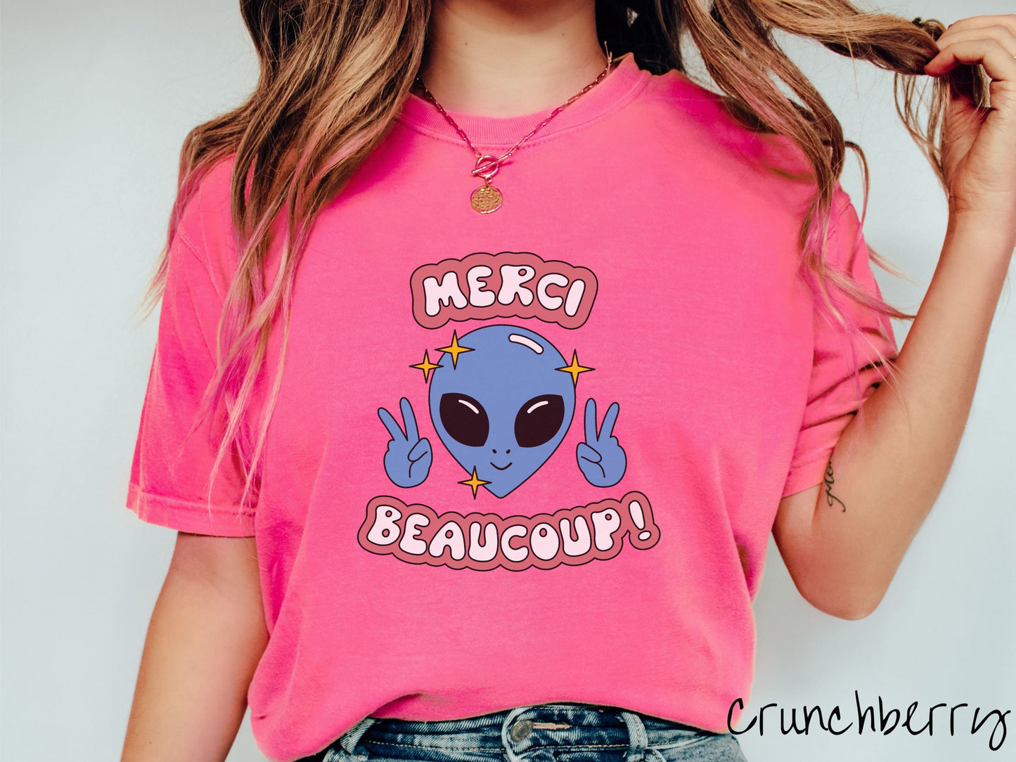 A woman wearing a cute, vintage crunchberry colored Comfort Colors shirt with the text Merci Beaucoup! in white and maroon colored font. In between that is a blue alien with large black eyes smiling and holding up two peace signs.