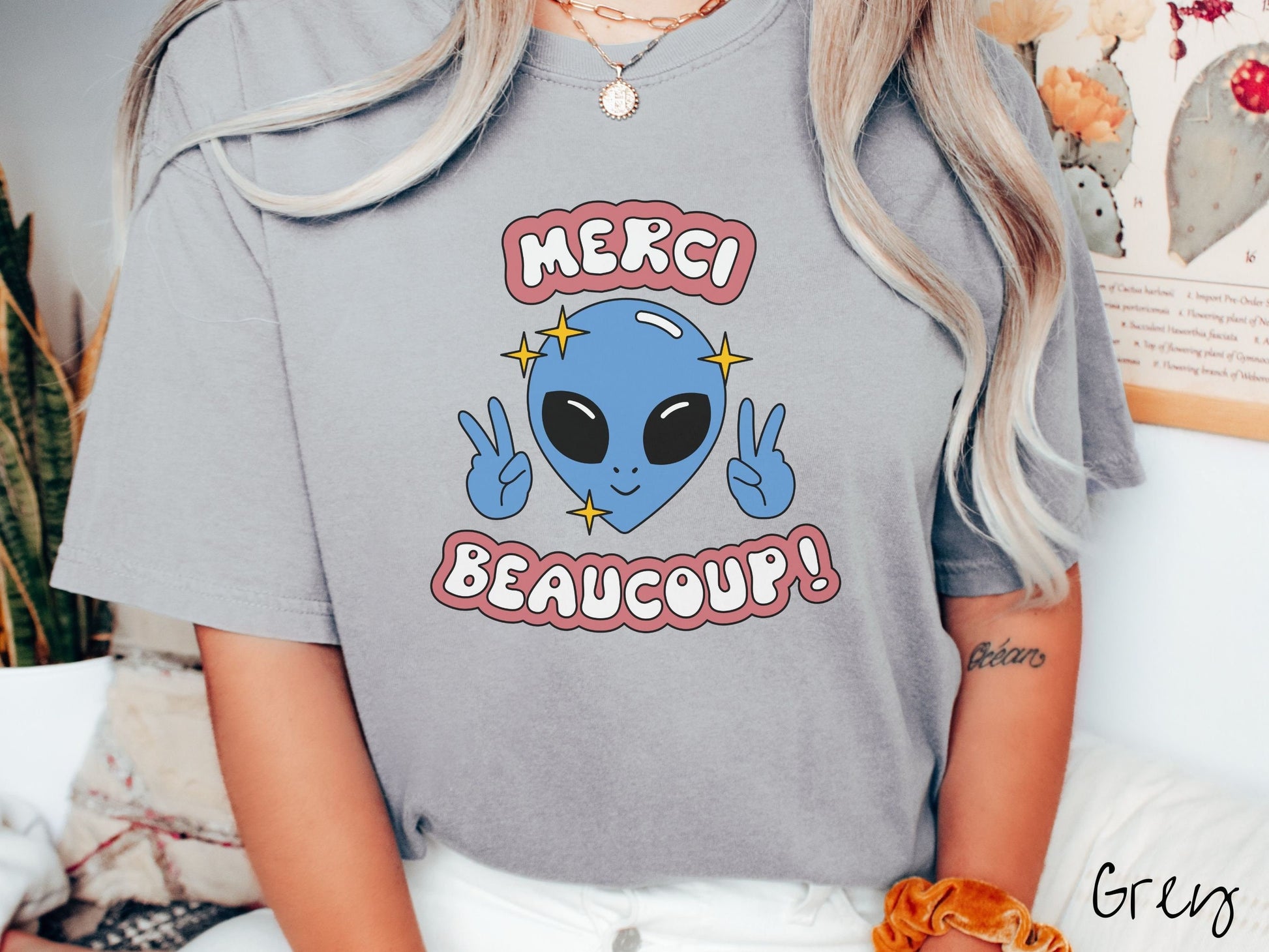 A woman wearing a cute, vintage grey colored Comfort Colors shirt with the text Merci Beaucoup! in white and maroon colored font. In between that is a blue alien with large black eyes smiling and holding up two peace signs.