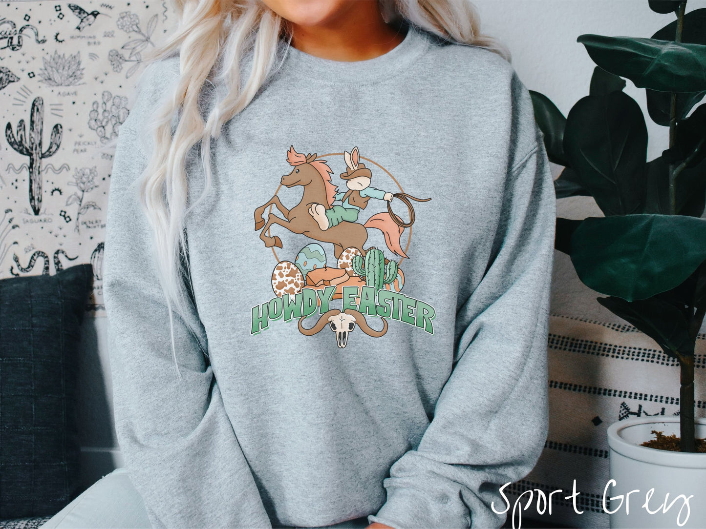 A woman wearing a cute, vintage sport grey colored sweatshirt with a cowboy rabbit riding a bucking, brown horse while holding a lasso rope. Below is green text Howdy Easter along with colorful Easter eggs, a green cactus, and a horned bull skull.