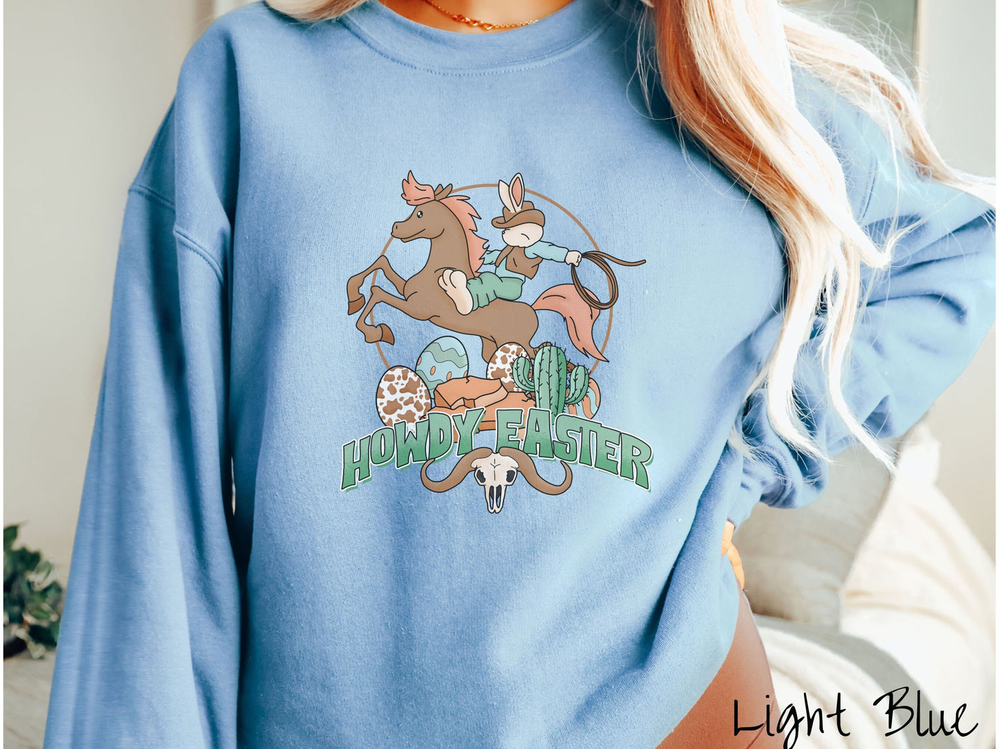 A woman wearing a cute, vintage light blue colored sweatshirt with a cowboy rabbit riding a bucking, brown horse while holding a lasso rope. Below is green text Howdy Easter along with colorful Easter eggs, a green cactus, and a horned bull skull.