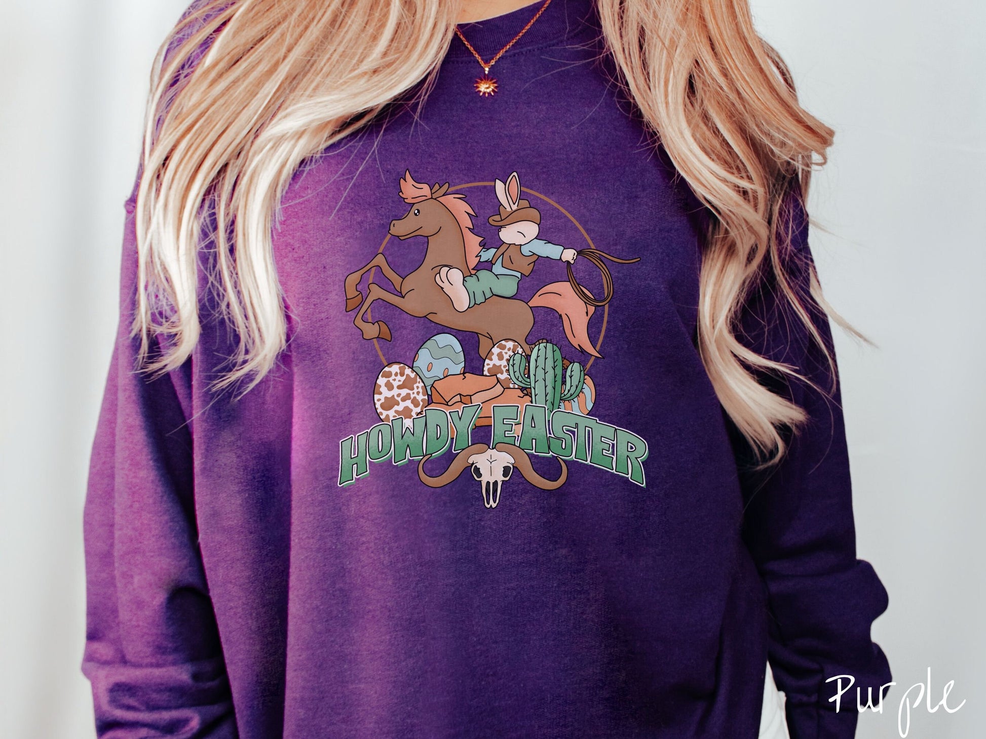 A woman wearing a cute, vintage purple colored sweatshirt with a cowboy rabbit riding a bucking, brown horse while holding a lasso rope. Below is green text Howdy Easter along with colorful Easter eggs, a green cactus, and a horned bull skull.