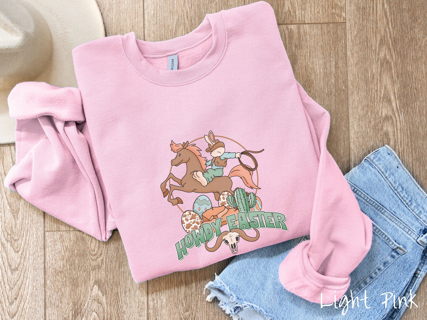 A cute, vintage light pink colored sweatshirt with a cowboy rabbit riding a bucking, brown horse while holding a lasso rope. Below is green text Howdy Easter along with colorful Easter eggs, a green cactus, and a horned bull skull.