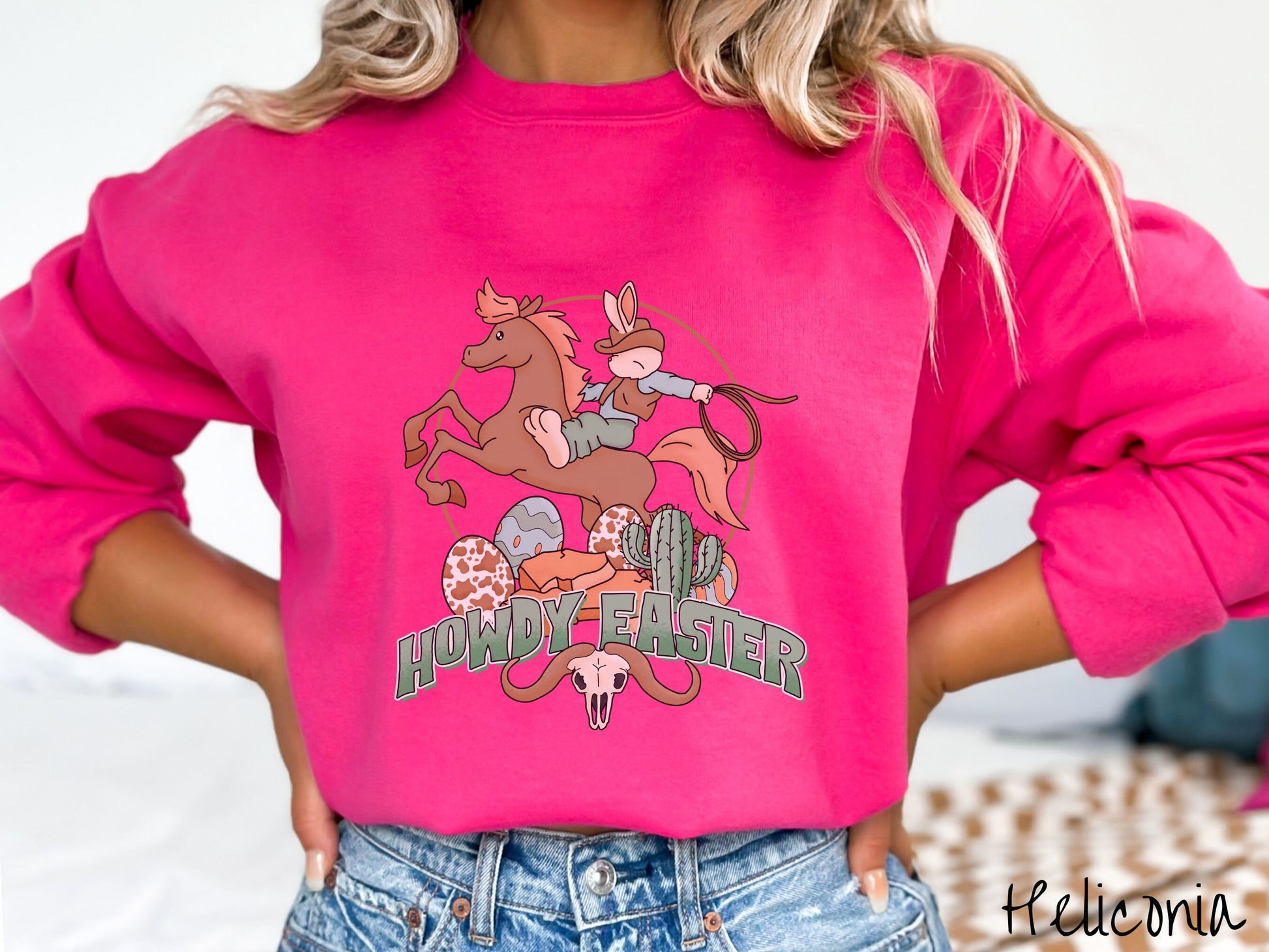 A woman wearing a cute, vintage heliconia colored sweatshirt with a cowboy rabbit riding a bucking, brown horse while holding a lasso rope. Below is green text Howdy Easter along with colorful Easter eggs, a green cactus, and a horned bull skull.