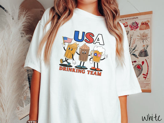 A woman wearing a cute, vintage white colored shirt with the letters USA in red, white, and blue font across the top. Below are three mugs of beer with hands and feet holding American flags, below them is the text Drinking Team in red font.