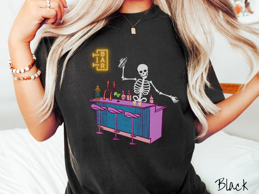 A woman wearing a cute, vintage black colored Comfort Colors t-shirt showing a skeleton bartender behind a purple and blue bar mixing drinks. There is a yellow bar sign behind the bar and three purple chairs in front. There are liquor bottles on top.