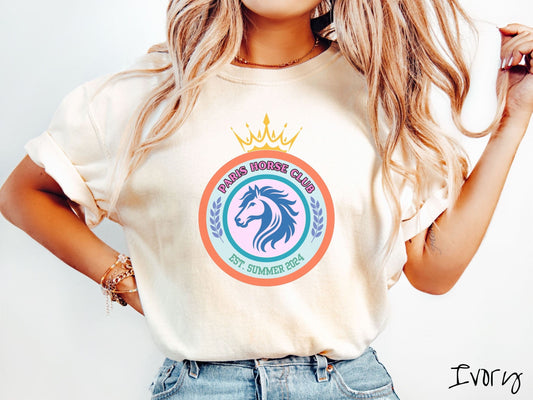 A woman wearing a cute, vintage ivory colored Comfort Colors shirt with a colorful coat of arms with a horse in the middle of two concentric circles, a crown on top. There is pink text along the top Paris Horse Club and below that Est. Summer 2024.