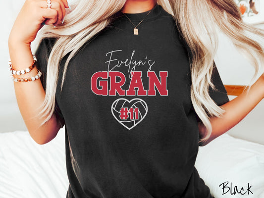 A woman wearing a cute black colored t-shirt with white and red text saying Evelyn&#39;s Gran. Below is a white, heart-shaped volleyball outline with #11 in red font written across it.